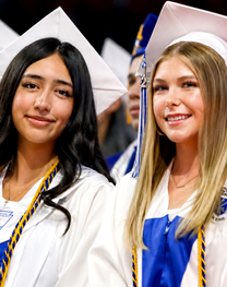 Two graduates wearing white caps and white-and-blue commencement gowns seated during a graduation ceremony.