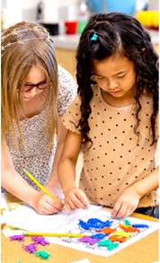 Two students working together at a table, drawing and arranging colorful craft materials on a large sheet of paper in a classroom setting.