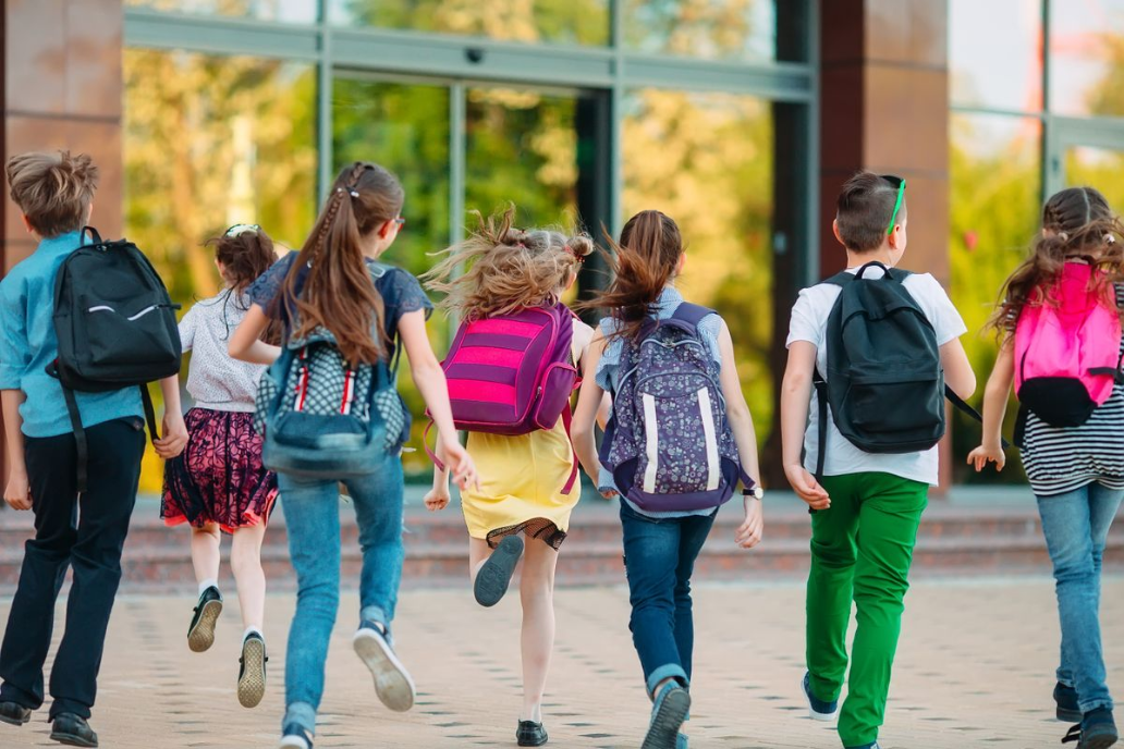 Kids with colorful backpacks joyfully run towards a school entrance. The scene conveys excitement and energy, with a bright, sunny atmosphere.